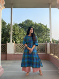 A woman stands barefoot on a covered outdoor balcony wearing a blue and green plaid kurta with pink wide-leg pants. Trees and a clear sky are visible in the background.