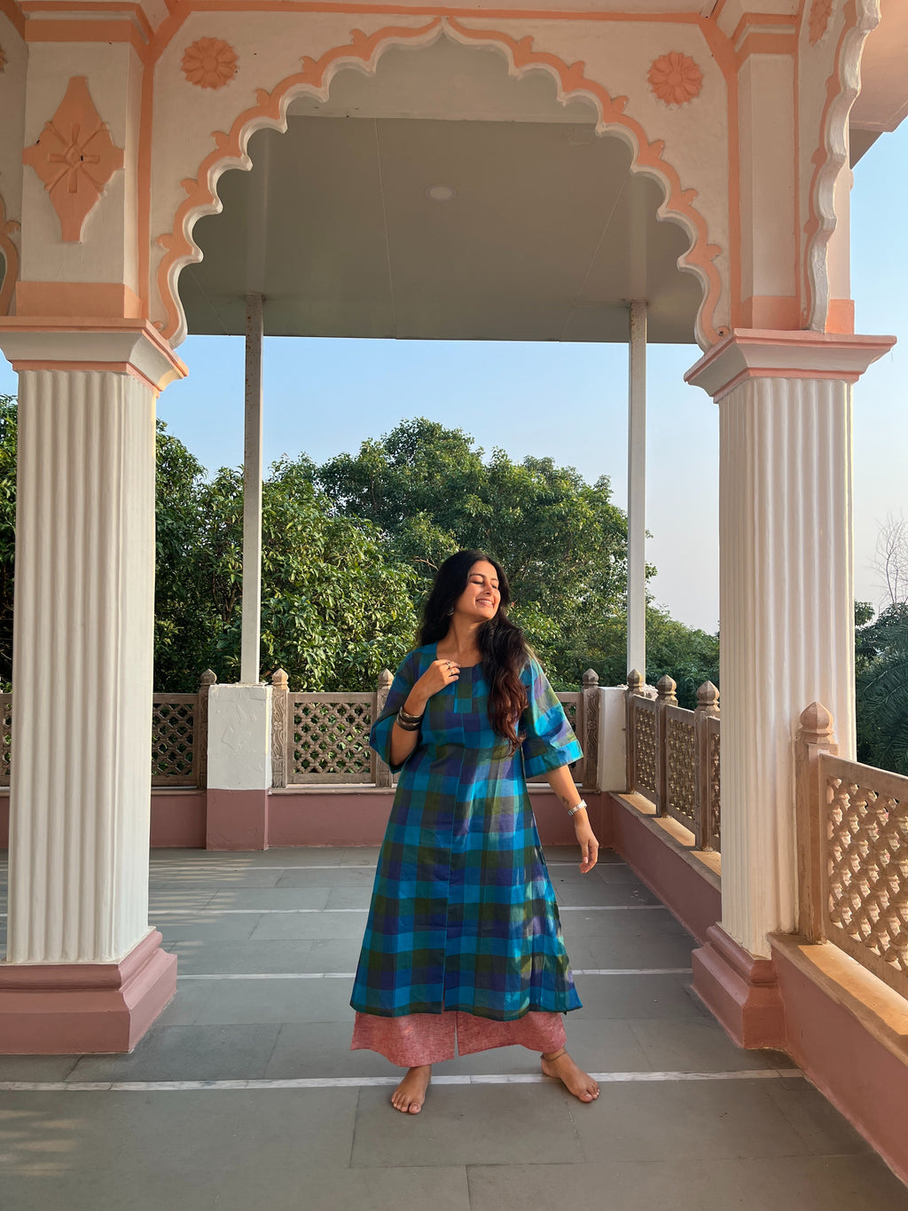 A woman stands barefoot in a covered walkway with pink and cream arches, wearing a blue and green plaid maxi dress over a pink bottom by Afeemi