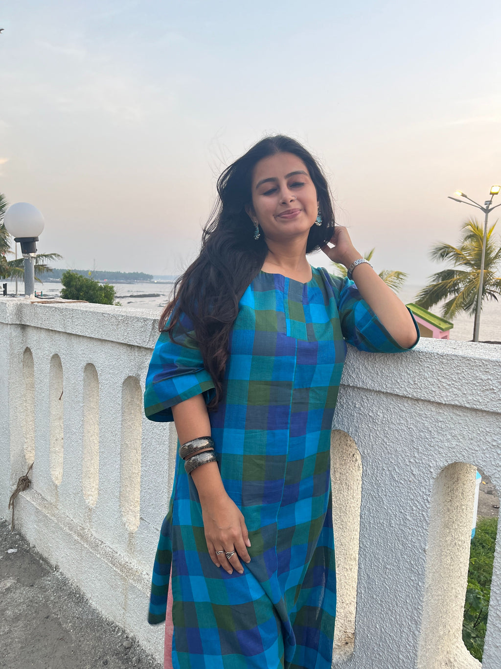 A woman wearing a blue and green plaid kurta stands on a white outdoor balcony railing with a scenic background.