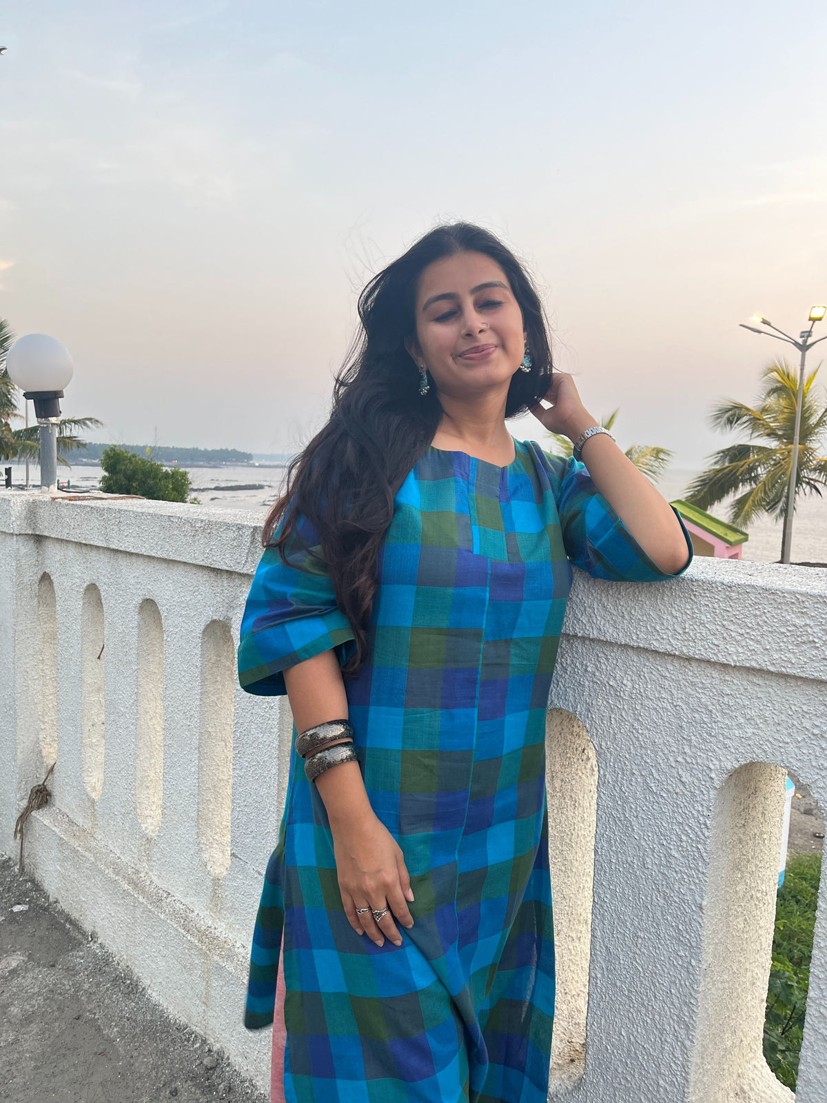 A woman wearing a blue and green plaid kurta stands on a white outdoor balcony railing with a scenic background.