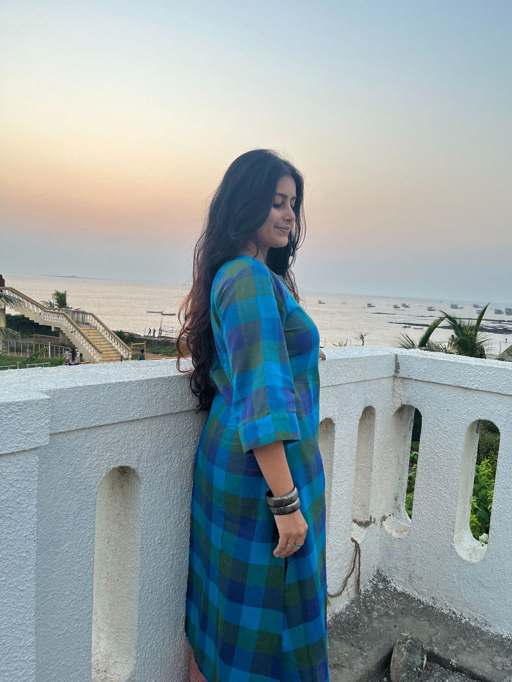 A woman wearing a blue and green plaid midi dress stands on a white balcony overlooking a coastal sunset and ocean.