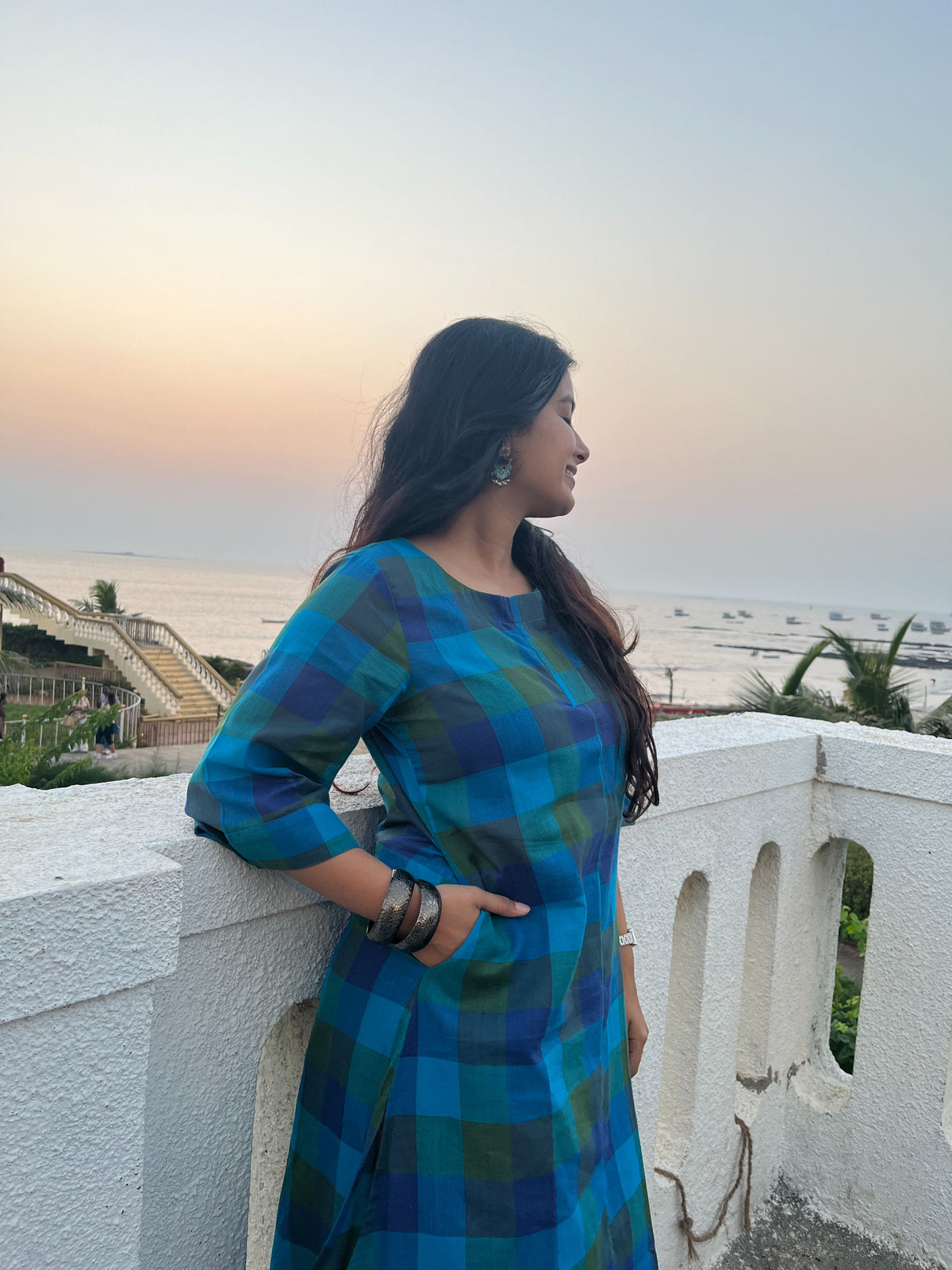 A woman wearing a blue and green plaid kurta stands in profile, leaning on a white stone wall with a beach and sunset in the background.