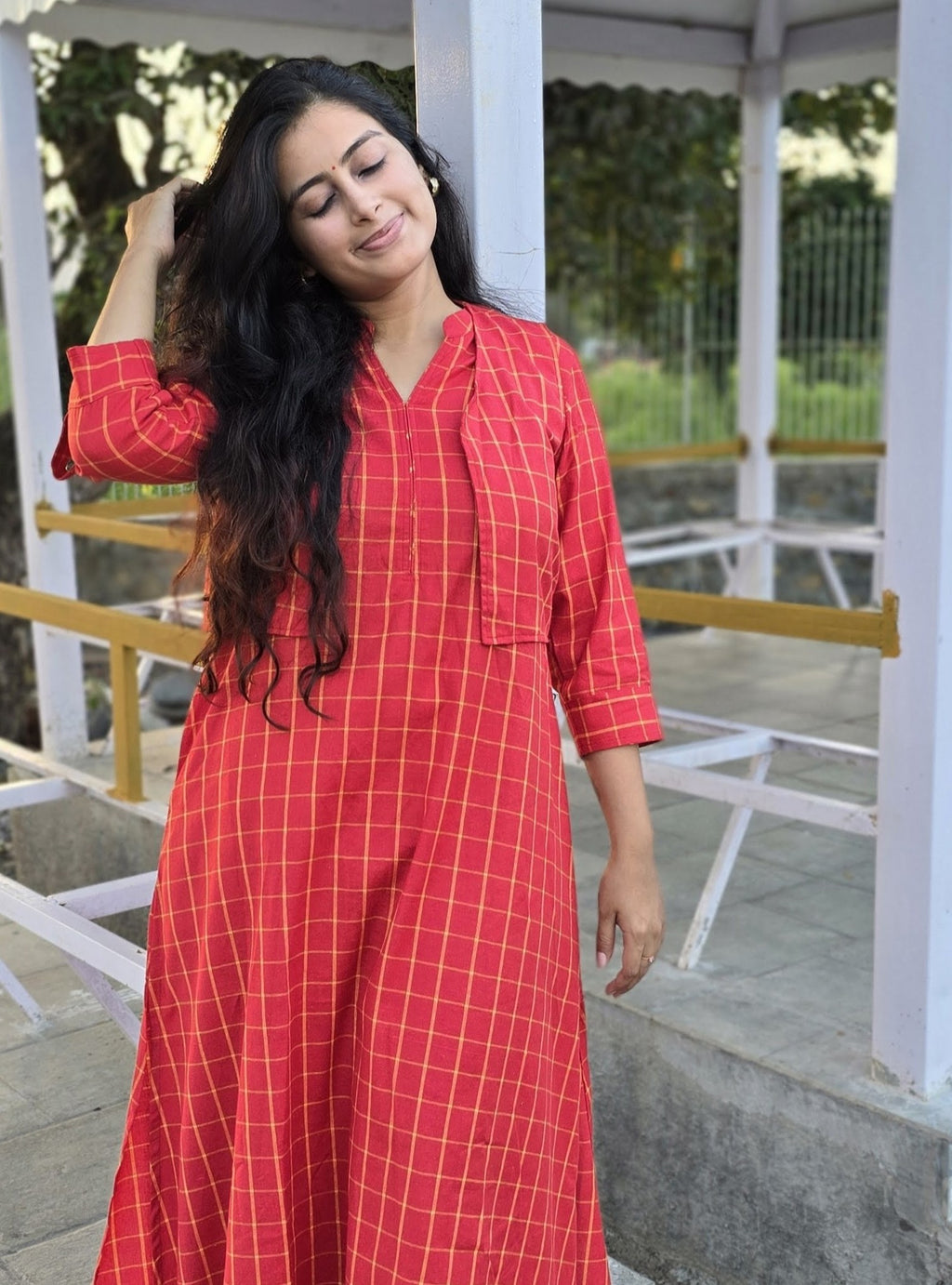 A woman wearing a red and yellow checkered long kurta standing in a white pavilion.